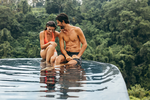 Happy young couple relaxing on the edge of outdoor swimming pool. Loving young man and woman enjoying holiday.
