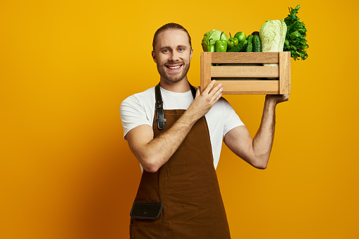 Happy young man in apron carrying wooden crate with fresh veggies against yellow background