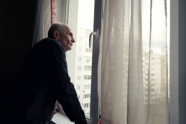 Grandfather looking through window glass covered with transparent white curtain. Senior man waiting guests with hope.