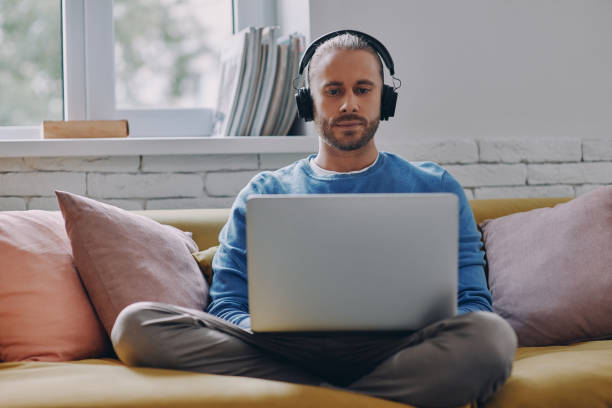 istock Confident young man in headphones working on laptop while sitting on the couch at home 1429971721