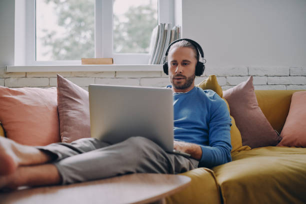 istock Concentrated young man in headphones working on laptop while sitting on the couch at home 1429972787