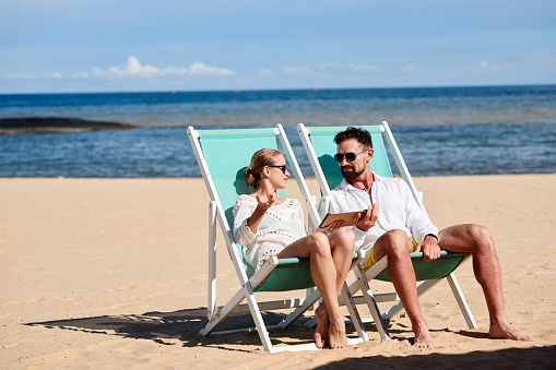 Young couple sitting on sunbeds and talking to each other using digital tablet on the beach