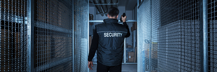 Rear View Of A Security Guard Standing In The Warehouse Holding Flashlight