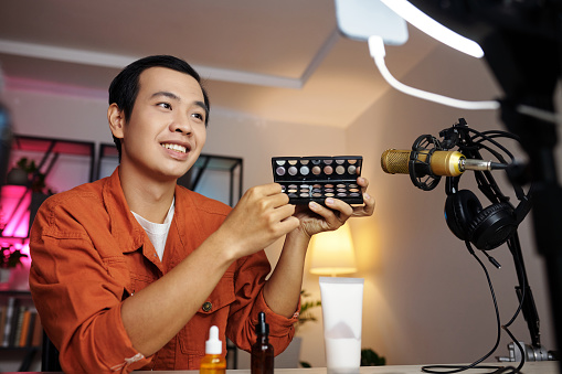 Horizontal medium portrait of cheerful young Asian man making video for blog demonstrating new eyeshadow palette on camera