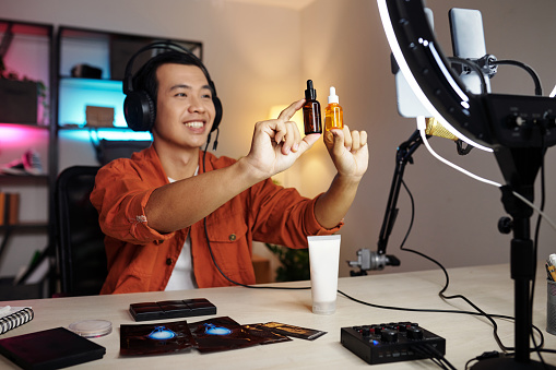 Selective focus medium portrait of joyful young Asian man sitting at desk in home studio making promotion video for new skincare products