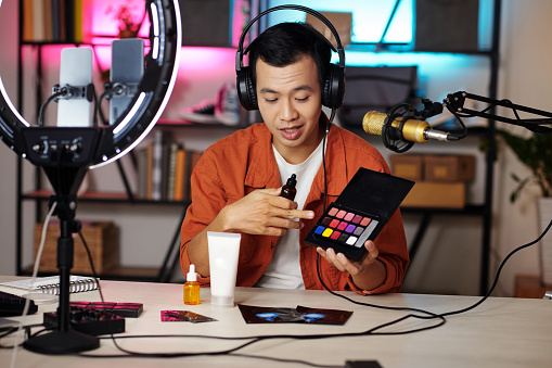 Modern Asian male influencer sitting at desk in home studio holding eyeshadow palette and face serum bottle making content for blog