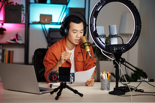Horizontal medium shot of handsome young Asian influencer wearing headphones sitting at desk in home studio recording podcast