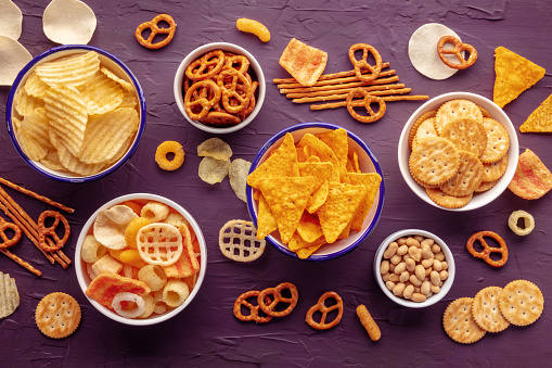 Salty snacks. Party food mix. Potato and tortilla chips, crackers and other appetizers in bowls, overhead flat lay shot on a vibrant purple background