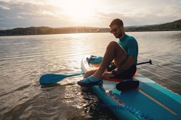 istock Caucasian man sitting on a standup paddleboard, while adjusting sports shoe 1429988021