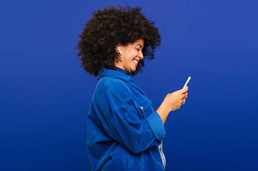Carefree young woman smiling happily while playing music using a smartphone and earbuds. Cheerful woman with curly hair enjoying her favourite playlist while standing against a blue background.