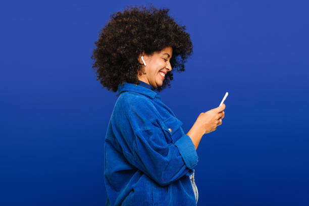 Carefree young woman playing music using a smartphone and earbuds Carefree young woman smiling happily while playing music using a smartphone and earbuds. Cheerful woman with curly hair enjoying her favourite playlist while standing against a blue background. cheerful stock pictures, royalty-free photos & images