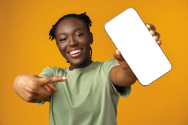 junge lächelnde afroamerikanerin zeigt smartphone mit leerem bildschirm vor gelbem hintergrund - luggage hold stock-fotos und bilder