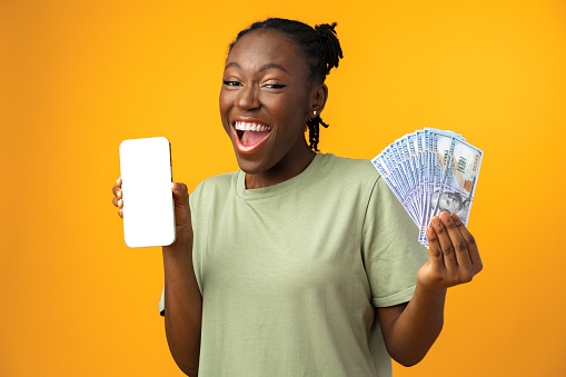 Young black woman holding white screen smartphone and money in yellow studio, close up