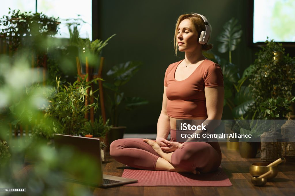 modern woman meditating in modern green house modern middle aged woman with yoga mat, laptop and headphones meditating in the modern green house. Meditating Stock Photo