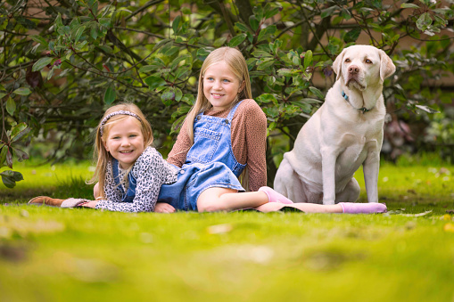 Siblings resting outdoors with their Labrador. Focus on older girl.
