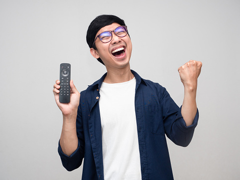 Cheerful positive asian man wear glasses show remote tv with fist up feels satisfied happy