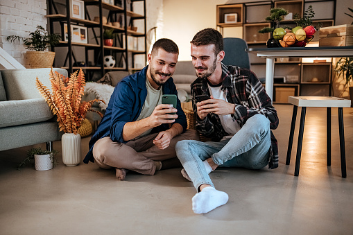 Happy gay couple using mobile phone while sitting on the floor