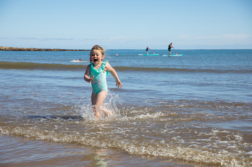 A young girl running out of the sea with a scared expression on her face in Northumberland wearing a blue swim suit.