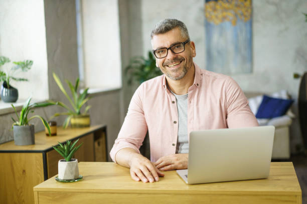 istock Handsome middle aged man in glasses working sitting at table with laptop at home. Freelancer man working at home using computer. Smiling man using laptop at home. Middle aged businessman at home 1430031792