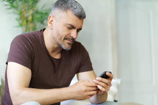 istock Middle aged handsome man holding smartphone texting or checking on social media, playing mobile game sitting on the couch in home interior. Grey haired man use phone sitting on the couch 1430031824