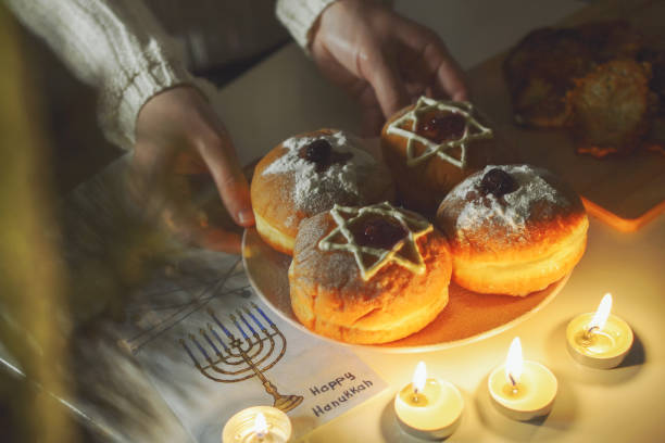 una vista dall'alto angolata del tavolo festivo di hanukkah, sul quale, accanto alle candele accese nel piatto a mano di una persona, vengono servite le tradizionali ciambelle sufganiya, decorate con la stella di david e zucchero a velo. - hanukkah menorah human hand lighting equipment foto e immagini stock