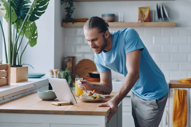istock Handsome man eating lunch and looking at the digital tablet while standing at the kitchen counter 1430033892