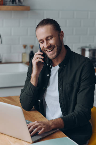 istock Cheerful young man talking on mobile phone and using laptop while sitting at the domestic kitchen 1430033958