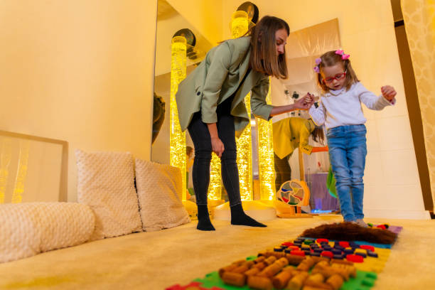 Therapist helping a young child living with cerebral palsy successfully improve her walking ability during therapy session in a sensory room, snoezelen. Texture walk, sensory exploration, motor skills. terapeuta que ayuda a un niño pequeño que vive con parálisis cerebral a mejorar con éxito su capacidad para caminar durante la sesión de terapia en una habitación sensorial, snoezelen. caminata de textura, exploración sensorial, habilidades motoras.
