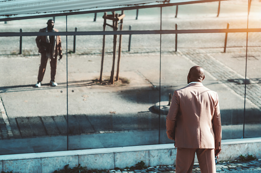 View from behind of an elegant bald black man in a pink tailored suit looking into a mirror wall facade of a street building which fully reflects him