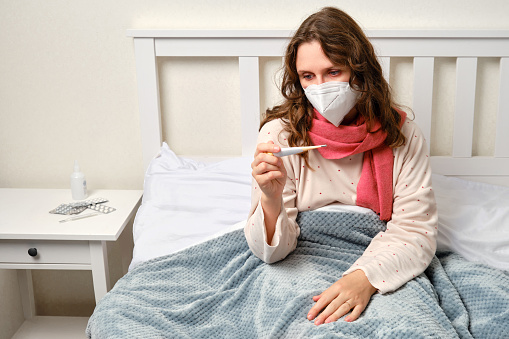 A sick woman is sitting on a bed in a medical face mask and a thermometer in her hands. Adult ill woman on a home bed in a white bedroom, female aged 35 years