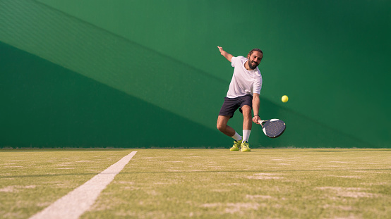 A close up view of a professional male tennis player running in mid motion reaching out above to strike the tennis ball in a volley from close to the baseline. The athlete is playing on a grass court in a generic stadium full of spectators. With motion blur.