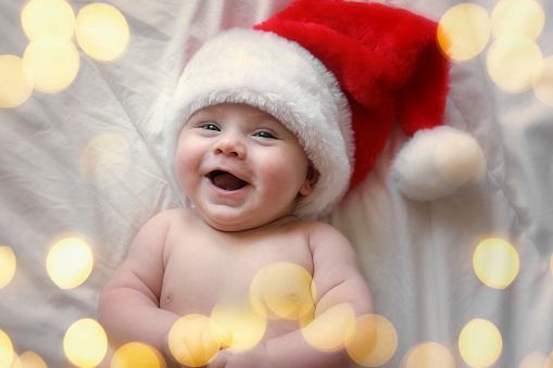 Smiling Baby Wearing Red Santa Claus Hat Celebrating Christmas. Cute Newborn Baby In Christmas Hat Lying On Knit Blanket. Merry xmas, Happy New Year, Infants, Holidays Concept. Funny Little Child. Christmas newborn baby. Little Toddler. Portrait of cute festive baby, wearing Santa hat, winter time. New Year. Cheerful kid smiling. Copyspace. Blurred Christmas lights on background.