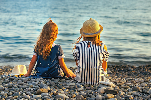 two little sister girls are sitting on the seashore at sunset. family travel during holidays
