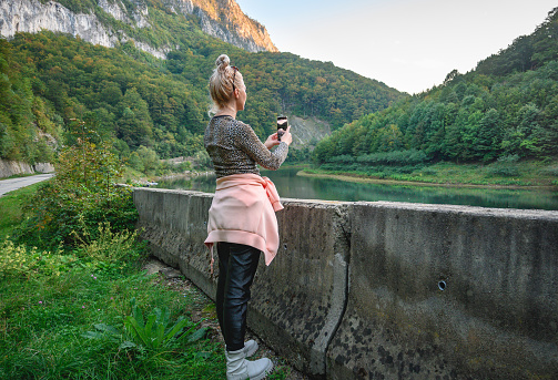 A woman with blond hair travels in a beautiful landscape and takes photos with her smartphone. Romania, Herculane. September, 29, 2022