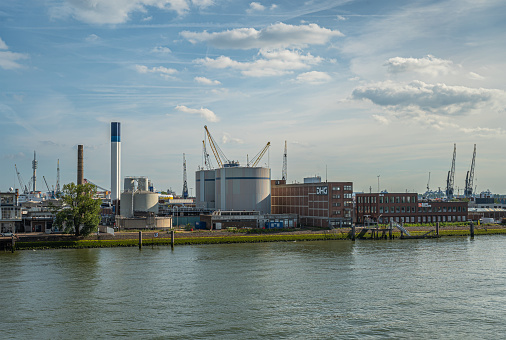 Rotterdam, Netherlands - July 11, 2022: Sludge processing platn Slusjesdijk with brown DHG offices along New Meuse under blue cloudscape. Industrial equipment and buildings on horizon