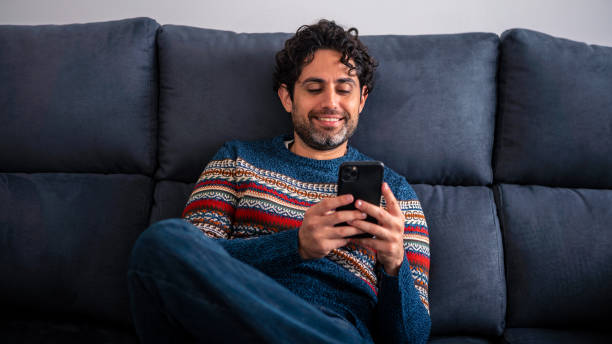istock Happy handsome man smiling while using smart phone and sitting on couch at home. 1430100877