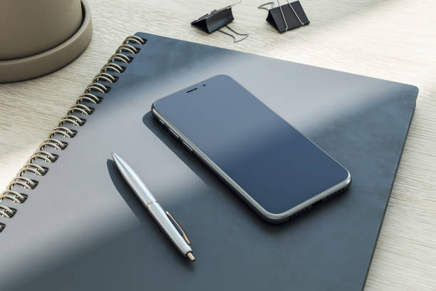 Top side view on blank dark smartphone screen with copyspace for your text or logo on black cover notebook with pen on abstract wooden surface. 3D rendering, mock up