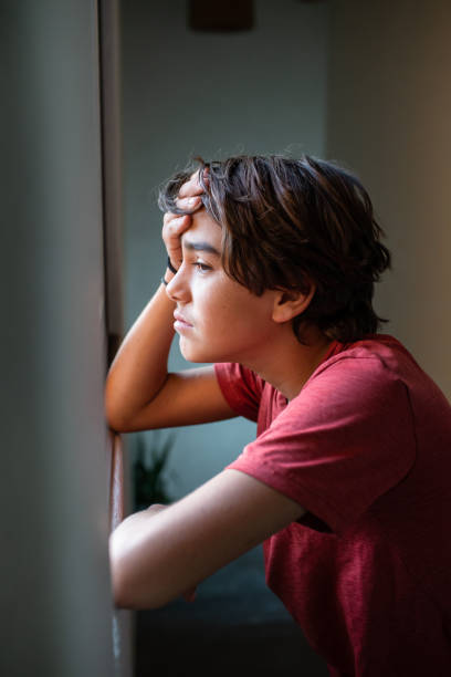 sad, worried pre adolescent boy looking out the window stock photo