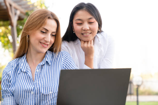 istock two young woman sitting in public park and using laptop 1430131828