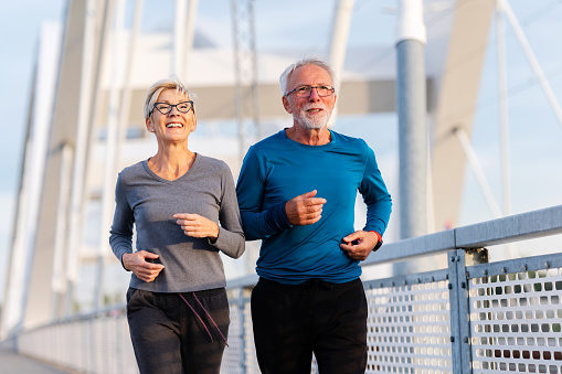 Cheerful active senior couple jogging together outdoors on the bridge. Healthy activities for elderly people.
