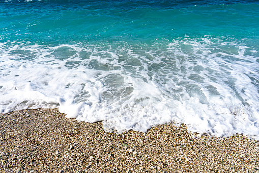 turquoise sea waves and white foam crashing on the sandy beach and small stones.
