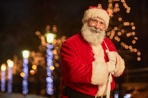 Waist up portrait of traditional Santa Claus holding sack with presents and smiling at camera in night city with lights, copy space