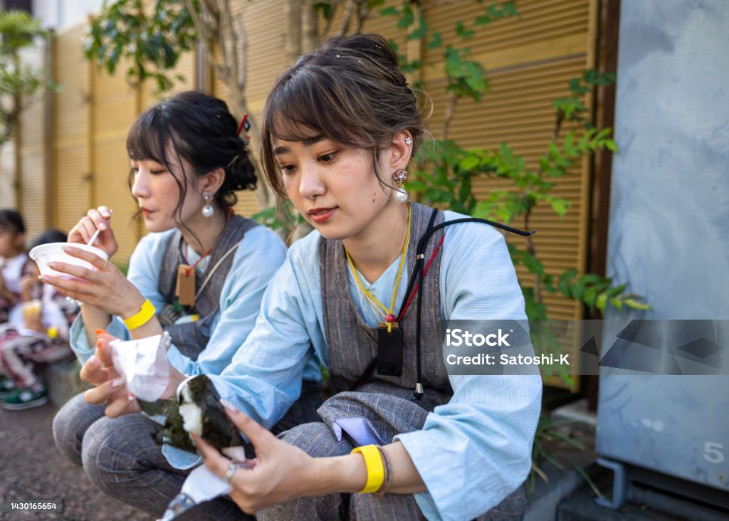 Young female Matsuri performers taking a break and eating 'Onigiri' rice ball on street Japanese people visiting traditional Japanese 'Matsuri' festival. Watching 'Matsuri' performance and joining their performance to pull a huge 'Dashi' float on street. Eating street foods, etc. 25-29 Years Stock Photo