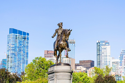 This bronze colored statue of George Washington is located at Boston  Public Garden in Massachusetts. In background of the image is skyscrapers of downtown Boston.