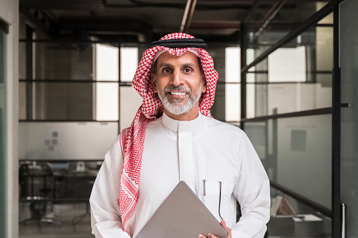 Waist-up front view of mature bearded man in traditional attire standing in modern office with portable information device and smiling at camera.