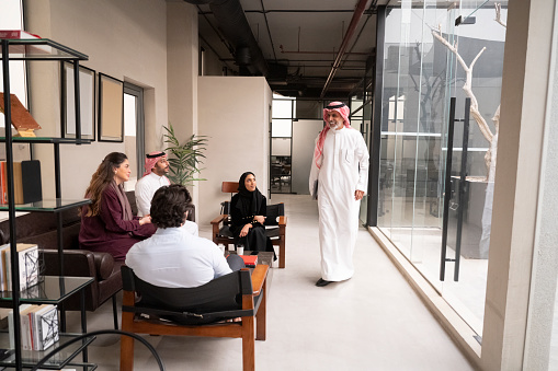 Full length view of Middle Eastern man in dish dash, kaffiyeh, and agal smiling at young project team gathered in office lounge area for informal update.