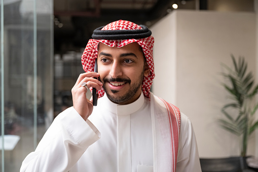 Head and shoulders view of smiling young executive with beard and mustache wearing traditional attire and looking away from camera while talking with caller.