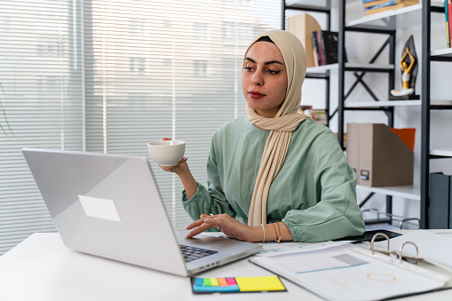 Businesswoman wearing hijab using desktop PC in office. Female professional is working at computer desk. She is wearing traditional clothing.