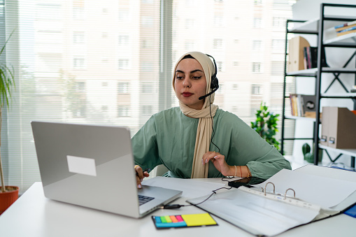 Muslim business woman with headset working in office. Happy arab woman working in company service center wearing headphone and using computer helping solving client problem. Islamic business woman working on computer at call center with copy space.