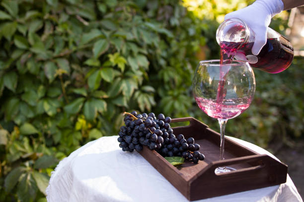A waiter's hand in a white glove pours a glass of red wine, which stands on a table with a white tablecloth outdoors in the sunlight stock photo